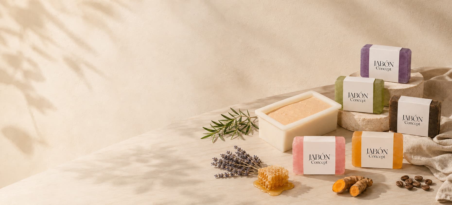 Năm viên xà bông quấn nhãn giấy trơn, đặt trên bệ đá travertine và mặt đá sáng màu; cạnh đó là khuôn xà bông vừa đổ, một nhánh lá xanh, bó oải hương khô, miếng sáp ong trên vũng mật, củ nghệ tươi và vài hạt cà phê. Nửa trái khung là mảng tường sáng với bóng lá lay động, bên phải là tấm vải lanh rủ xuống
