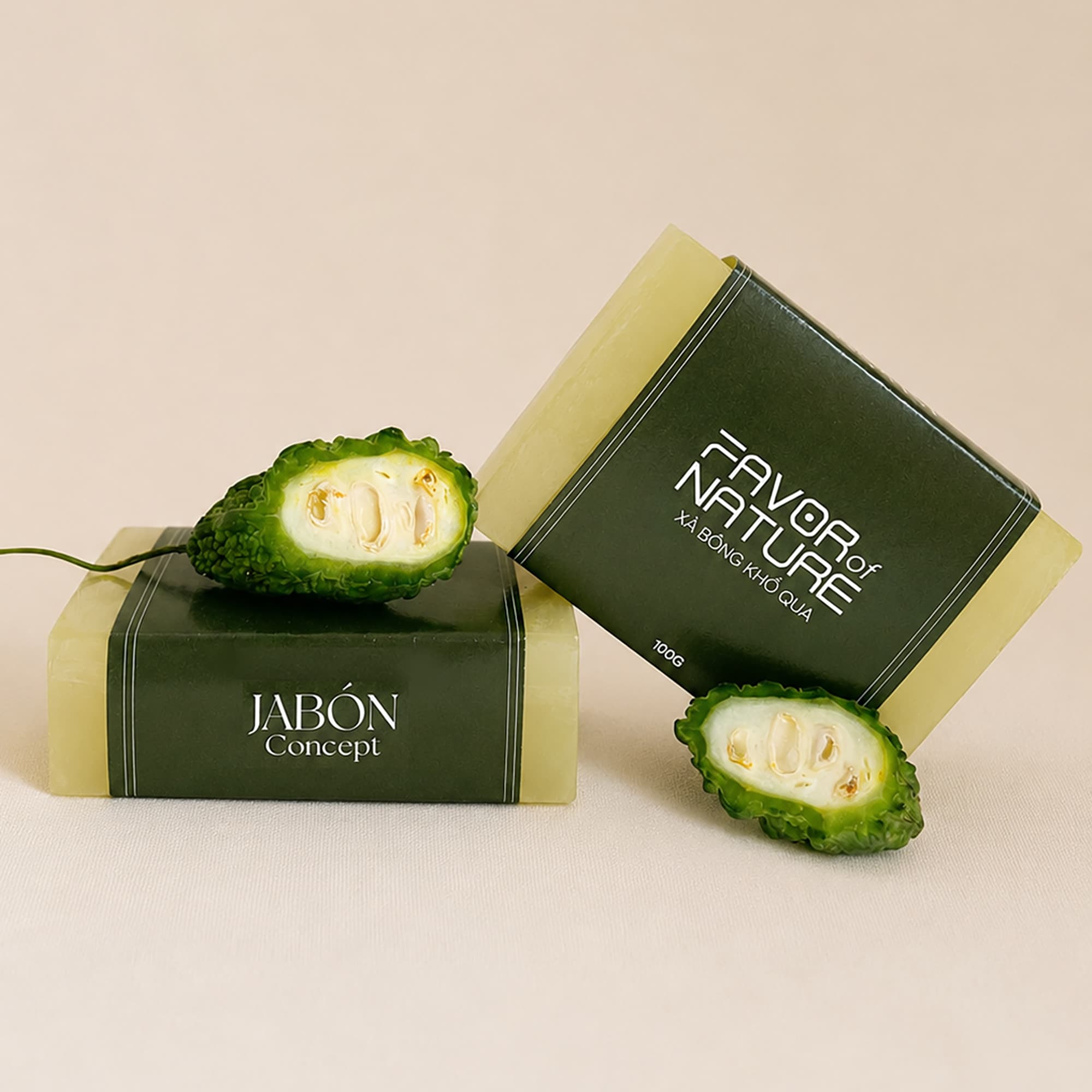 Hai viên xà bông khổ qua màu vàng xanh trong, quấn dải nhãn giấy xanh ô liu FAVOR of NATURE và JABÓN Concept, điểm hai nửa quả mướp đắng tươi