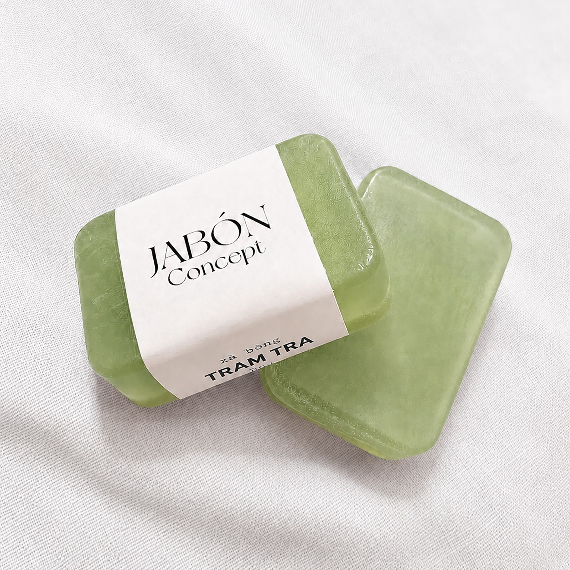 Hai viên xà bông tràm trà màu xanh lá nhạt, viên phía trước quấn dải nhãn giấy trắng Jabón Concept, đặt trên nền vải dệt trắng