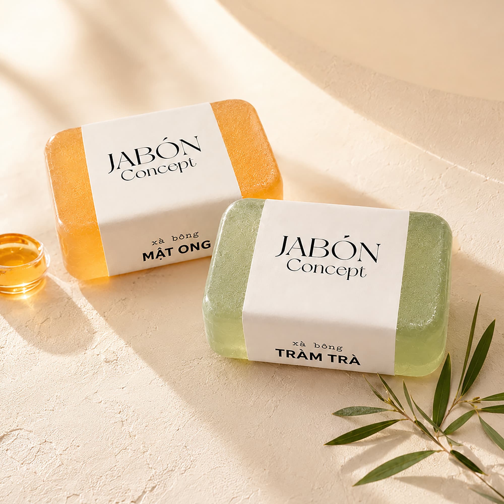 Hai viên xà bông Jabón Concept — một viên mật ong màu hổ phách, một viên tràm trà màu xanh nhạt — quấn dải nhãn giấy trắng, đặt cạnh chén mật ong và nhánh tràm trà