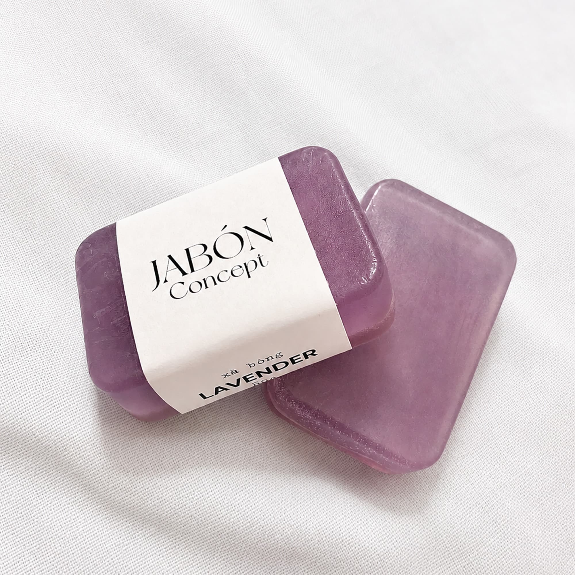 Hai viên xà bông lavender màu tím, viên phía trước quấn dải nhãn giấy trắng Jabón Concept, đặt trên nền vải dệt trắng